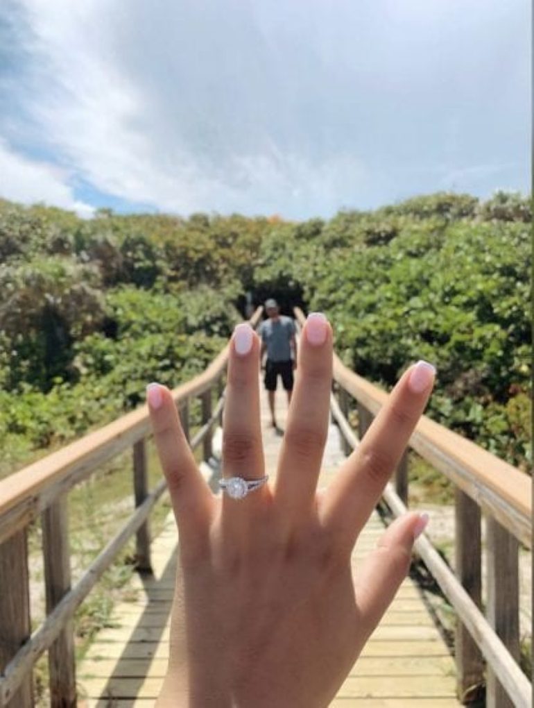 A woman shows off her new engagement ring on a bridge with her fianc&eacute; in the background.